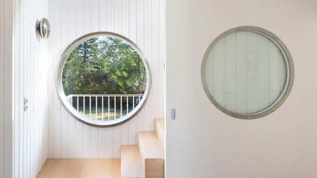 Ein runder Fensterblick auf Bäume und eine Holztreppe in einem hellen Raum.