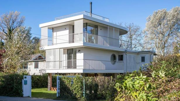 Ein modernes, zweistöckiges Haus mit Balkonen und einem Flachdach, umgeben von Bäumen und Büschen.