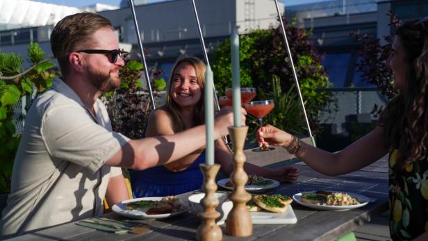 Auf dem Bild sind drei Personen abgebildet, die im Freien sitzen, mit einem Drink anstoßen und gegrillte Speisen genießen.