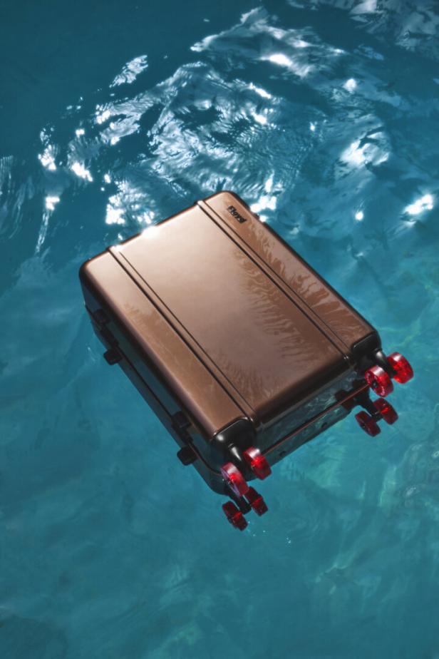 Ein bronzefarbener Rollkoffer treibt auf dem türkisfarbenen Wasser.