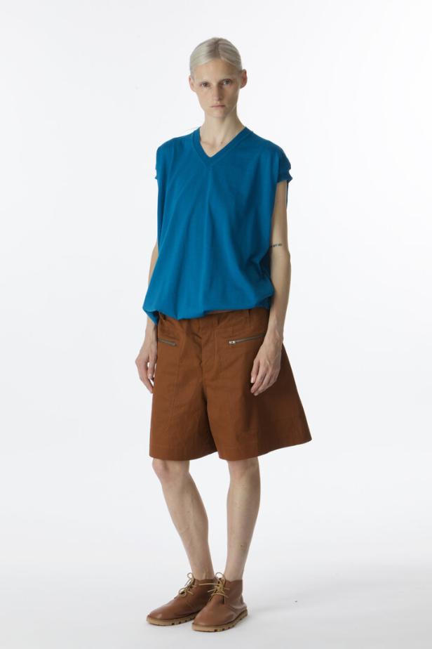 Eine Frau mit blonden Haaren trägt ein blaues T-Shirt, braune Shorts und braune Schuhe.