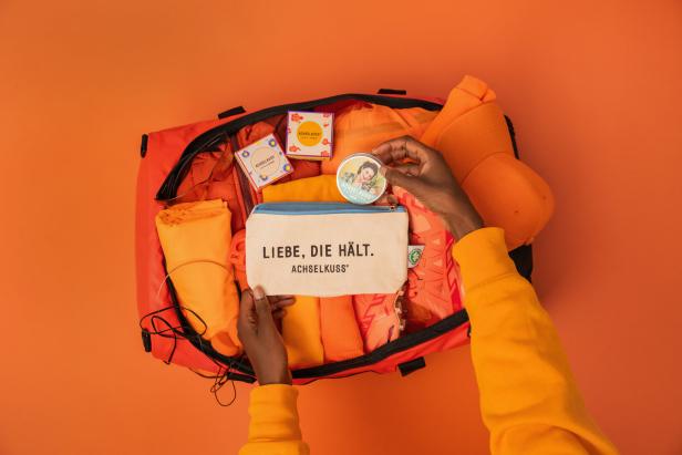 Eine Person packt eine Tasche mit Produkten der Marke „Achselkuss“ auf einem orangefarbenen Hintergrund.