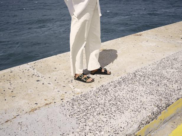 Eine Person in Sandalen und Leinenhose steht an einem Kai am Meer.