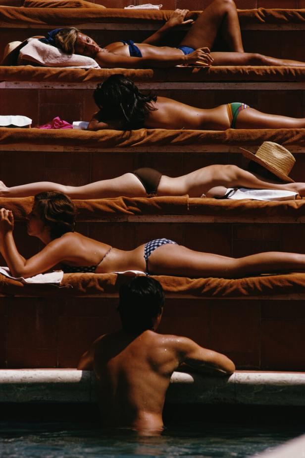 Sonnenbadende Frauen liegen auf Stufen. Ein Mann im Pool sieht zu ihnen hinauf.
