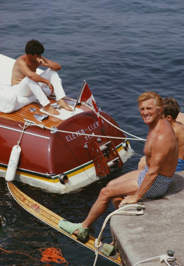 Kirk Douglas in Badehose sitzt an der Hafenmauer, er hat Wasserski an, vor ihm ein Motorboot.