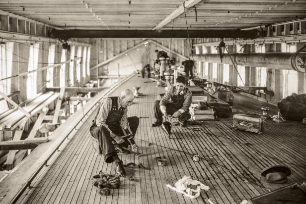 Zwei Männer arbeiten auf dem Deck eines Schiffes, möglicherweise beim Abdichten der Planken.