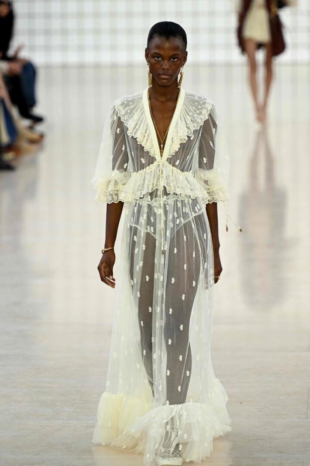 Eine Frau präsentiert ein transparentes, weißes Kleid mit Blumenmuster auf dem Laufsteg.