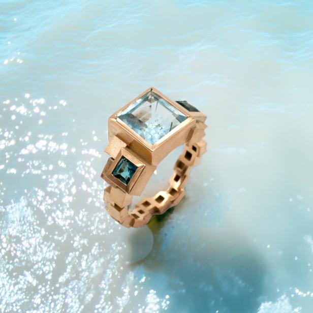 Ein goldener Ring mit einem großen, hellblauen und zwei kleineren, dunkelblauen Steinen liegt im Wasser.