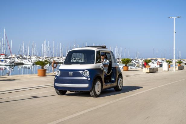 Ein blauer Fiat Topolino fährt an einem sonnigen Tag an einem Yachthafen entlang.