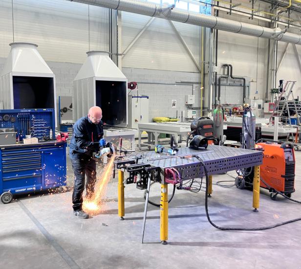 Ein Mann arbeitet mit einer Schleifmaschine an einem Metalltisch in einer Werkshalle.