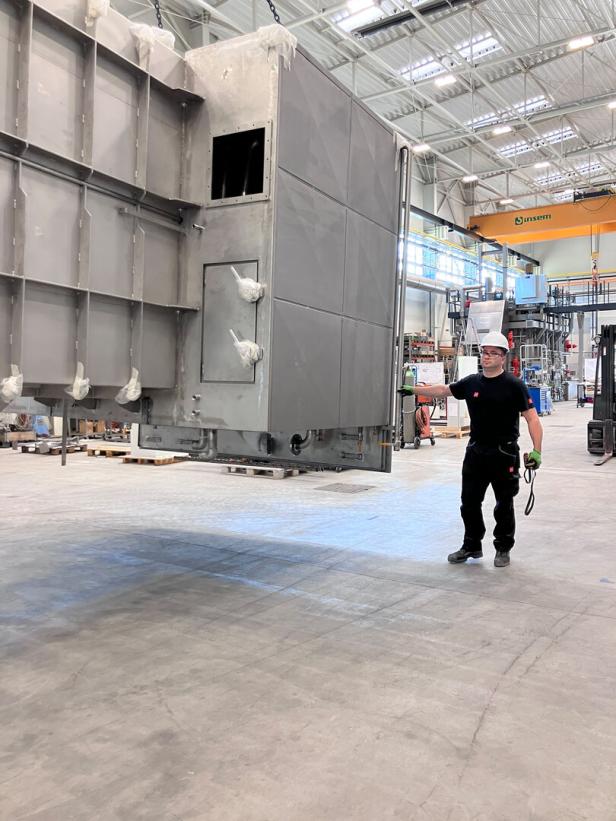 Ein Arbeiter mit Schutzhelm steht vor einer großen, grauen Industrieanlage in einer Fabrikhalle.