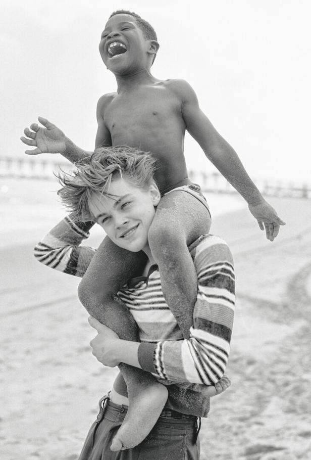 Ein junger Leonardo DiCaprio trägt einen lachenden Jungen huckepack am Strand.