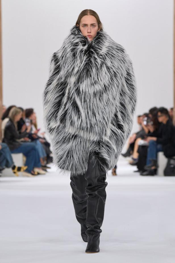 Ein Model auf dem Laufsteg trägt einen übergroßen Pelzmantel und schwarze Stiefel.