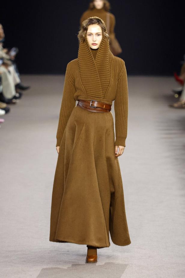 Ein Model präsentiert ein braunes Strickoberteil mit Rollkragen und einen langen, ausgestellten Rock von Max Mara.