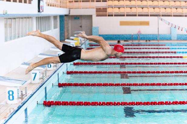 At start. Sportive male swimmer in red cap and goggles in motion and action during training at pool, indoors. Healthy lifestyle, power, energy, sports movement concept