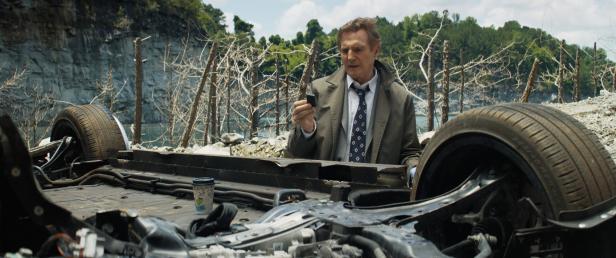 Liam Neeson steht an einem Autowrack und betrachtet ein kleines Gerät.