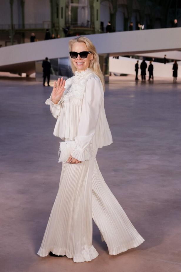 Eine Frau in einem weißen Hosenanzug mit Sonnenbrille winkt auf einer Chanel-Modenschau.