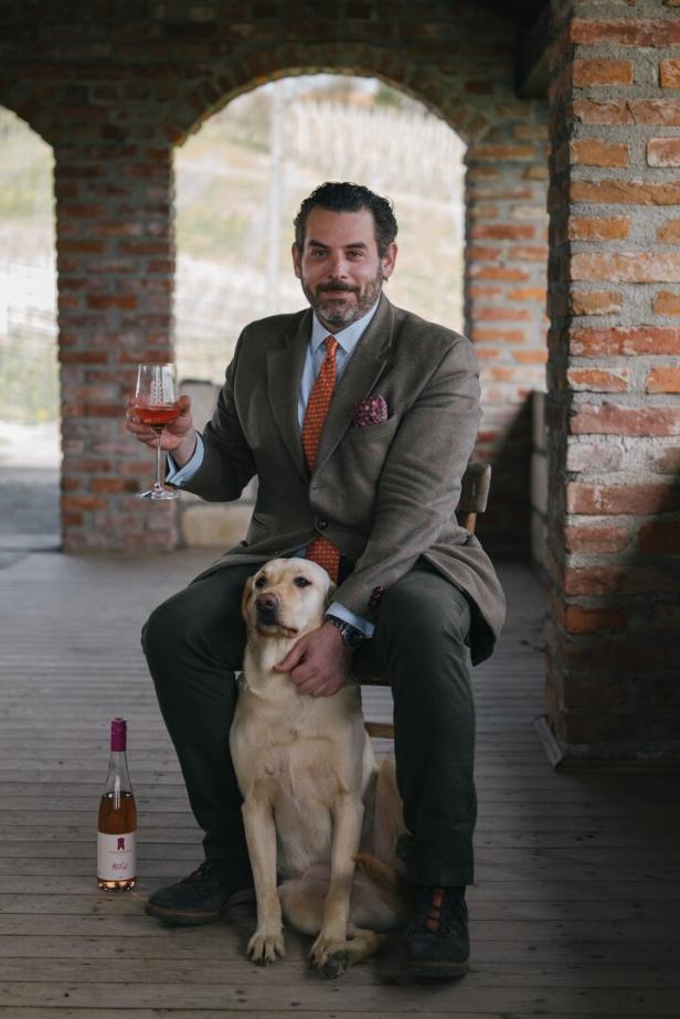 Ein Mann im Anzug sitzt mit einem Glas Rosé und einem Labrador vor einem Backsteingebäude.