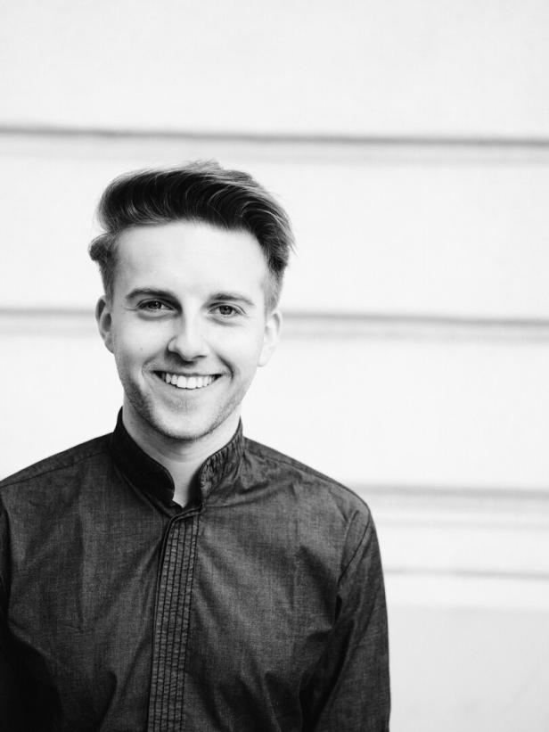 Ein lächelnder junger Mann in einem dunklen Hemd vor einer hellen Wand.