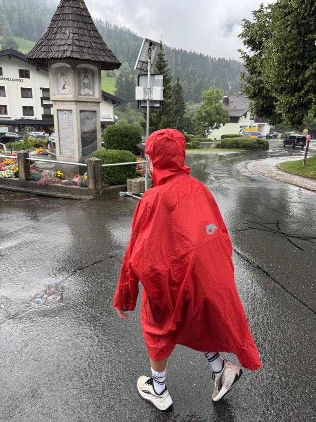 Eine Person in einem roten Regenponcho geht bei Regen durch ein Dorf.