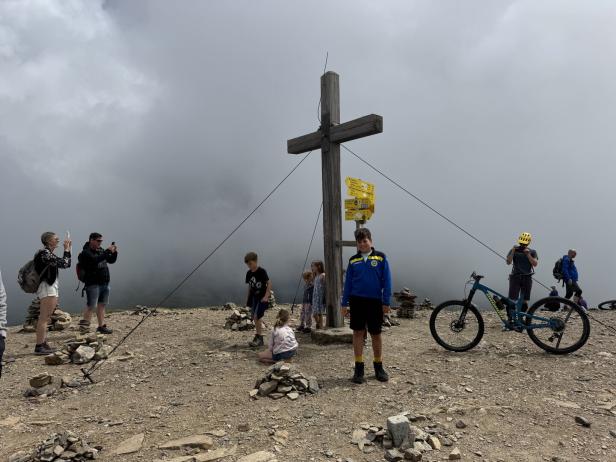 Eine Gruppe von Menschen steht auf einem Berggipfel mit Gipfelkreuz im Nebel.