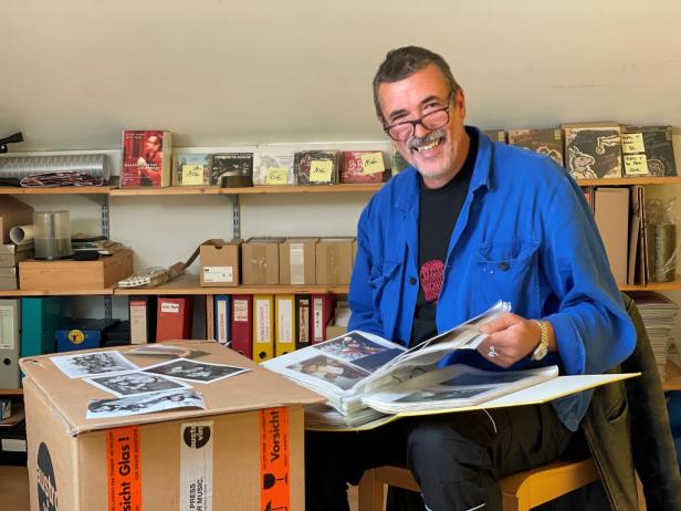 Ein Mann mit Brille sitzt in einem Raum voller Schallplatten und Ordner und blättert in einem Fotoalbum.