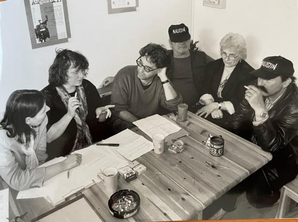 Eine Gruppe von Menschen sitzt an einem Tisch und diskutiert, einige tragen „Augustin“-Kappen.