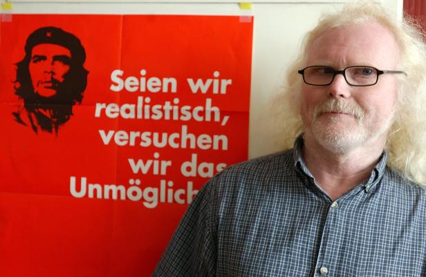 Ein Mann mit Brille steht vor einem roten Poster mit einem Bild von Che Guevara und dem Zitat „Seien wir realistisch, versuchen wir das Unmögliche“.