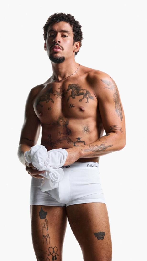 Ein tätowierter Mann mit Unterwäsche von Calvin Klein hält ein weißes T-Shirt.
