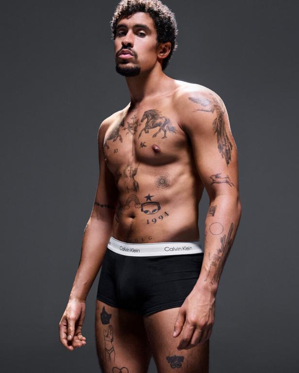 Ein Mann mit Tattoos und Calvin Klein Unterwäsche posiert vor einem grauen Hintergrund.