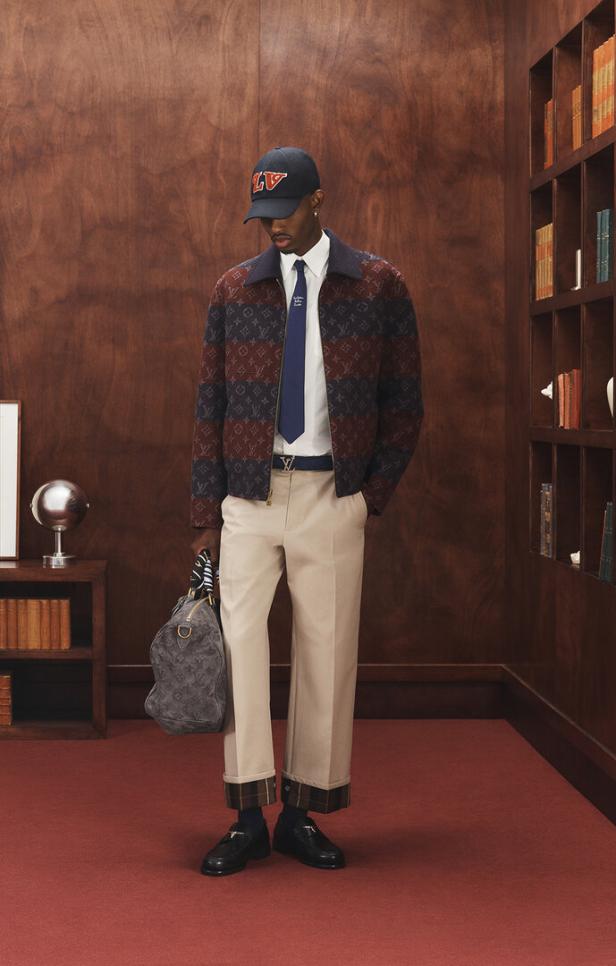 Ein Mann in einer Louis Vuitton Jacke, Hose und Kappe posiert in einem Raum mit Bücherregalen.