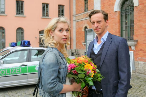 Eine Frau mit Blumenstrauß und ein Mann stehen vor einem Polizeiauto und einem Gebäude mit Backsteinfassade.