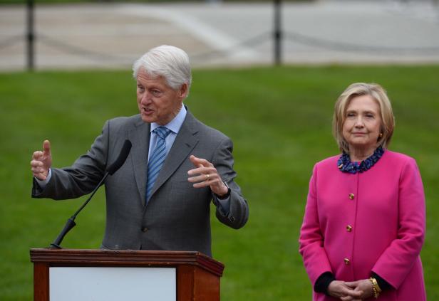 Bill Clinton spricht an einem Rednerpult, während Hillary Clinton daneben steht.