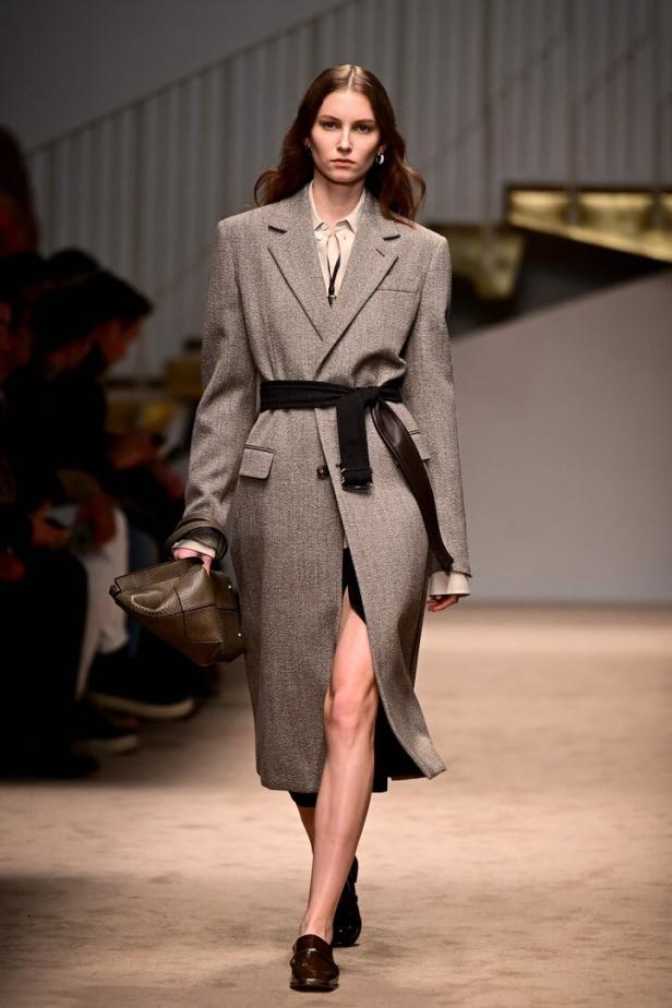 Ein Model präsentiert einen langen, grau-braunen Mantel mit Gürtel auf dem Laufsteg.