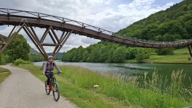 Eine Frau fährt mit dem Fahrrad unter einer ungewöhnlichen Brücke entlang eines Flusses.