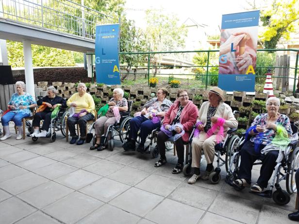 Eine Gruppe älterer Damen sitzt im Freien, einige im Rollstuhl, und hält bunte Tücher.