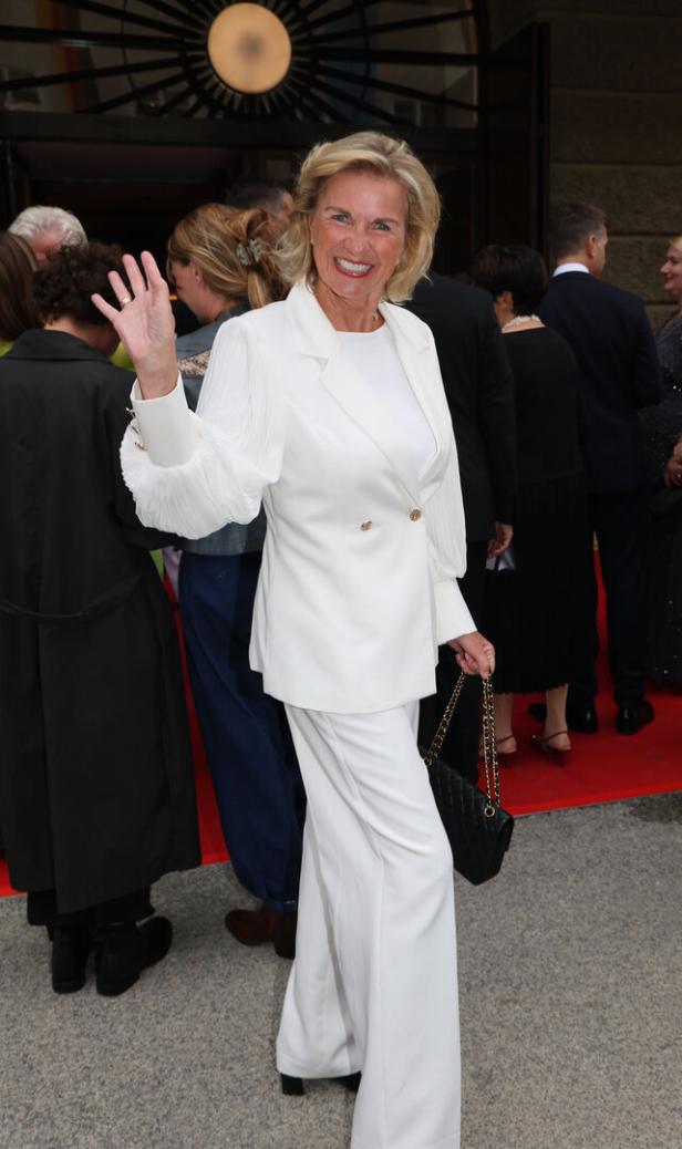 Eine Frau in einem weißen Hosenanzug winkt auf einem roten Teppich.
