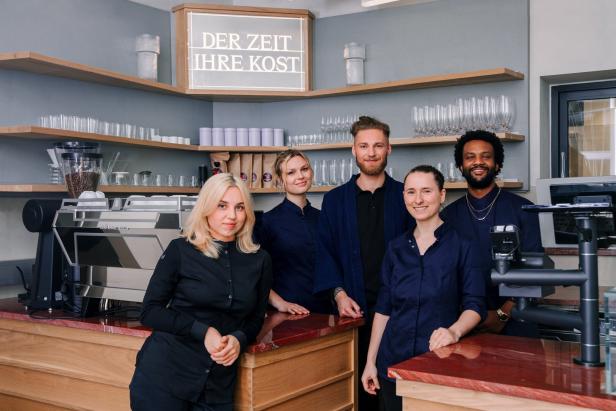 Das Team eines Cafés steht hinter der Theke unter einem Schild mit der Aufschrift „Der Zeit ihre Kost“.