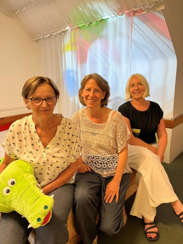 Drei Frauen sitzen nebeneinander, eine hält ein grünes Krokodil-Kuscheltier.