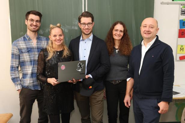 Auf dem Bild sind fünf Personen abgebildet, die vor einer Tafel in einer Schulklasse stehen und in die Kamera lächeln.