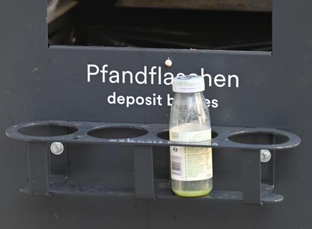 Eine leere Pfandflasche steht in einer Halterung an einem Mülleimer.