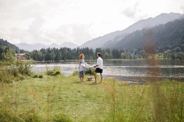 Ein Paar breitet am Ufer eines Bergsees die Picknickdecke aus, umgeben von majestätischen Bergen.