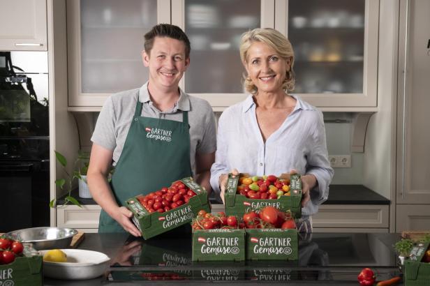 Vor einer Küchenzeile halten zwei Personen Kisten mit Tomaten und lächeln in die Kamera.