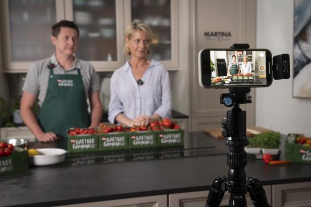 Zwei Personen stehen in einer Küche vor vier Kisten Tomaten und einer Kamera.