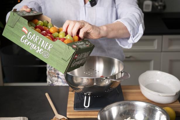 Eine Person gibt Tomaten aus einer Kiste in einen Sieb, der auf einer Küchenwaage steht.