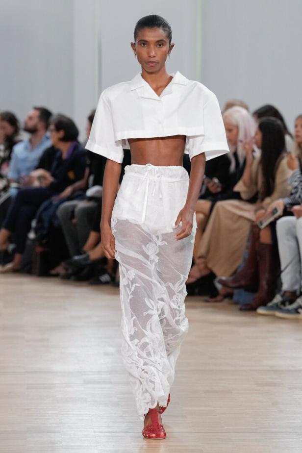 Eine Frau präsentiert ein weißes Outfit mit transparenten, floralen Elementen auf einem Laufsteg.
