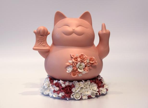 Eine Winkekatze aus Porzellan steht auf Blumen, ist mit Blumen geschmückt, zeigt aber den Mittelfinger.