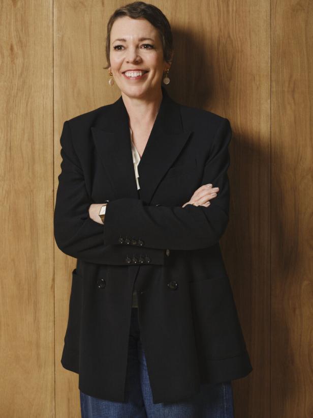Olivia Colman lächelt mit verschränkten Armen vor einer Holzwand.