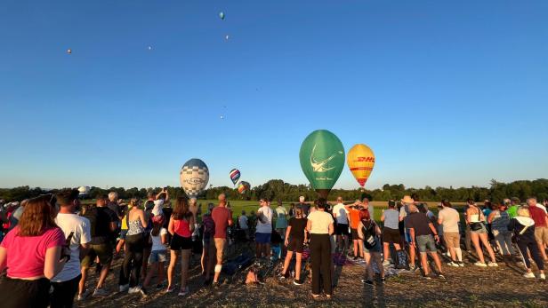 Zahlreiche Menschen stehen auf einer Wiese und schauen Richtung Horizont, wo erste Heißluftballone zu sehen sind.