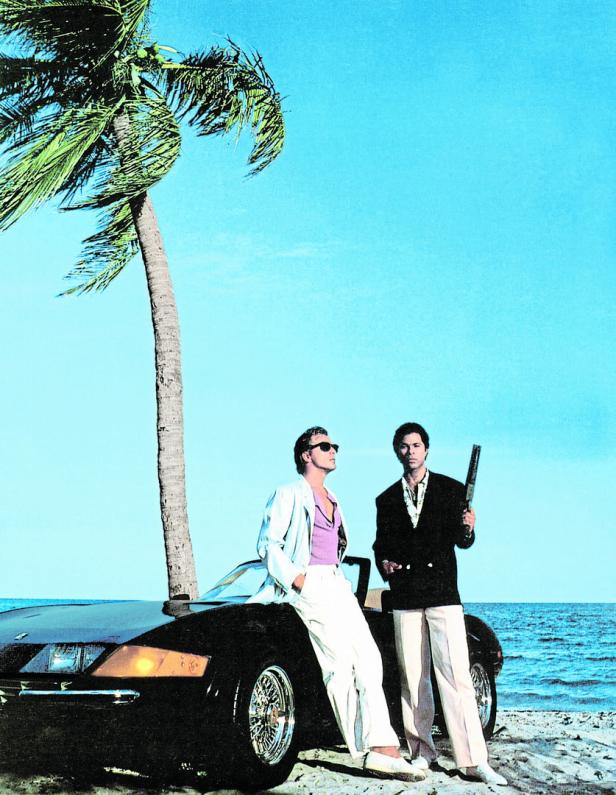 Zwei Männer im Stil von „Miami Vice“ posieren mit einem Sportwagen am Strand.